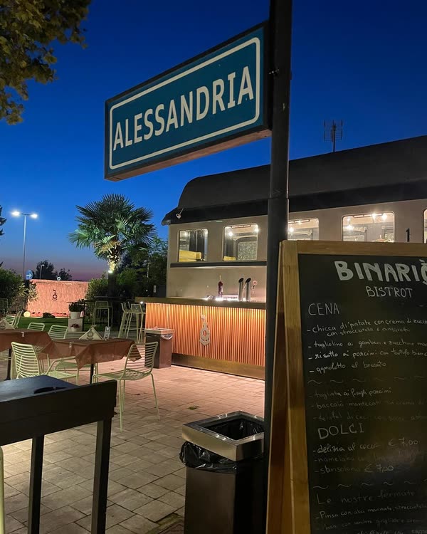 Il cartello ALESSANDRIA all'ora blu, con il bancone esterno e la lavagna del menu sotto la carrozza
